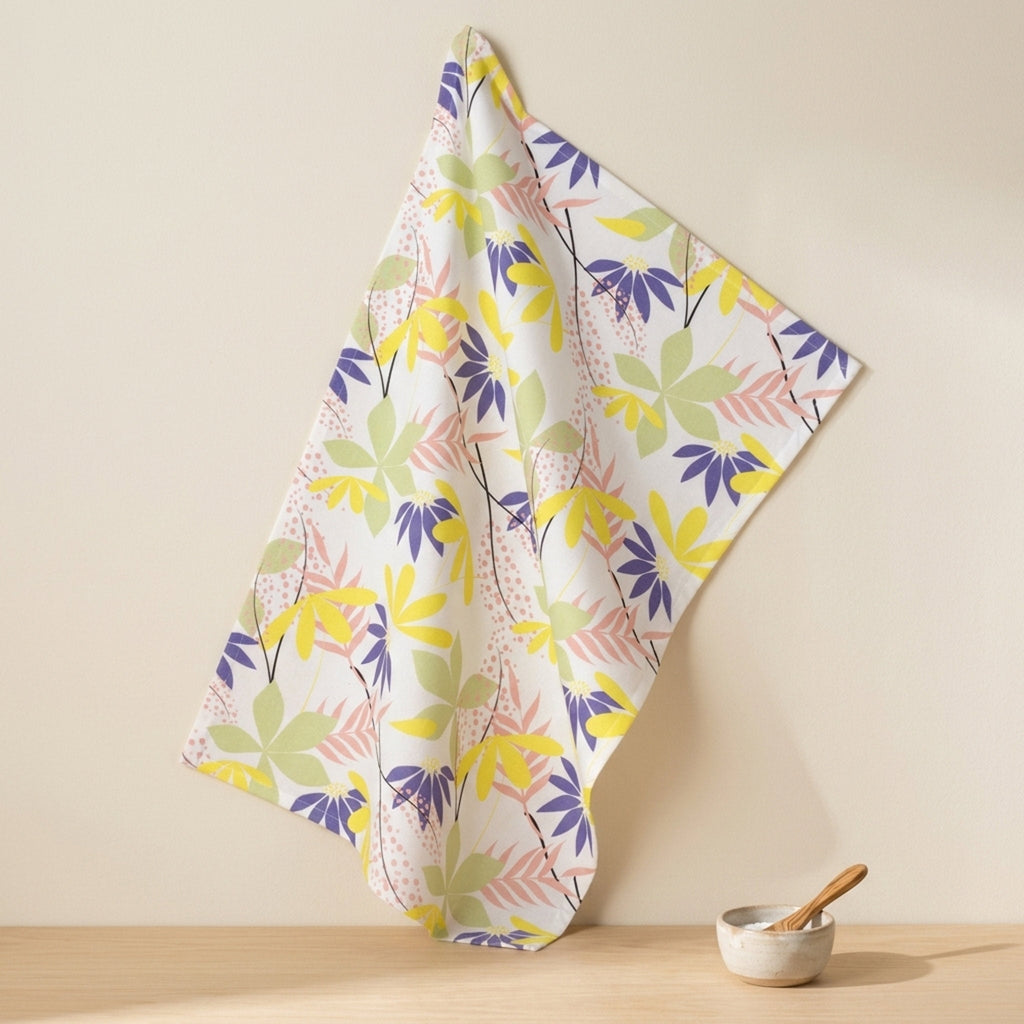 A cotton kitchen towel with a colorful botanical leaf pattern in yellow, purple, green, and pink on a white background, hanging against a beige wall above a wooden surface with a small bowl and spoon from Hem And Tassel