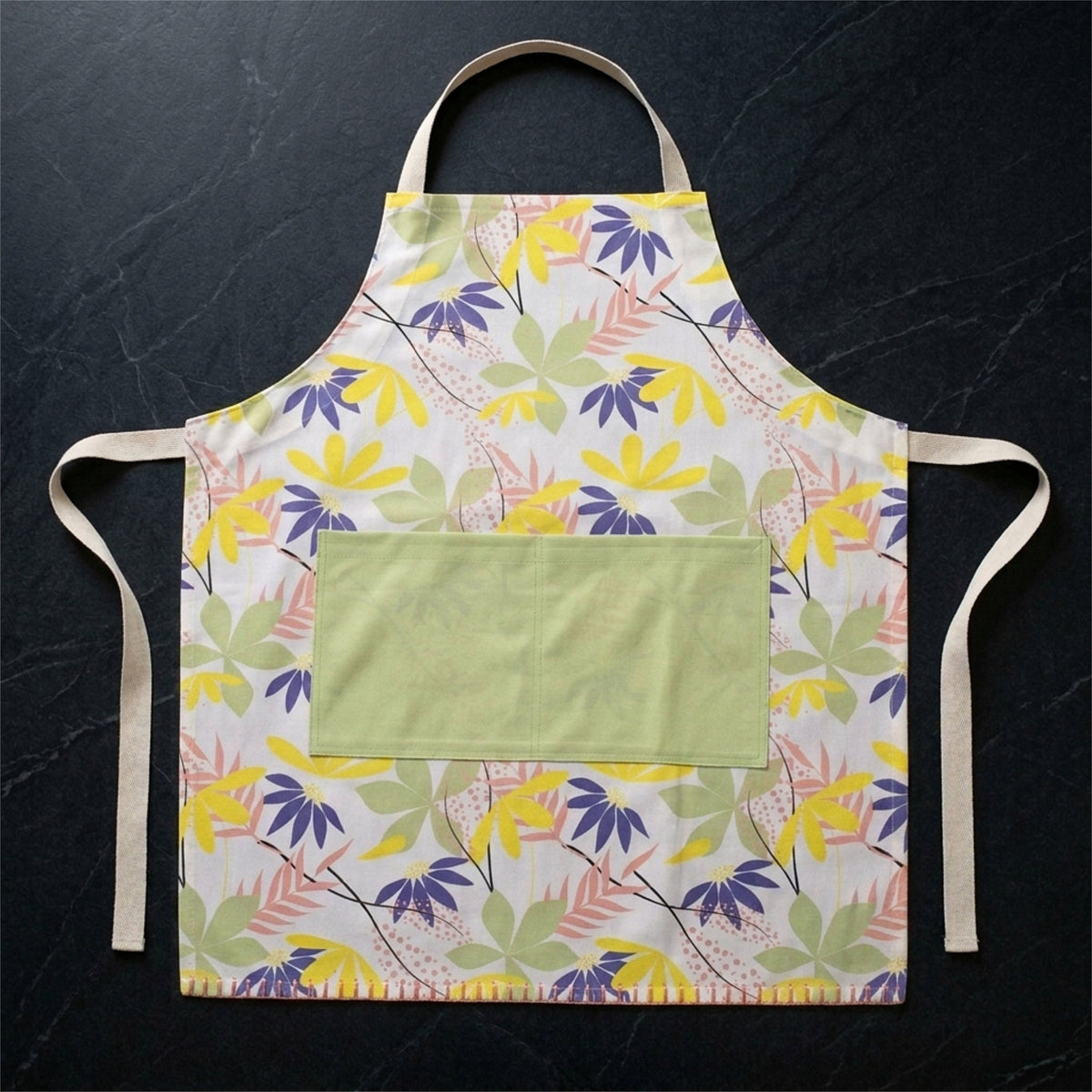 A floral apron with a light green front pocket, laid flat on a dark textured background. The apron features a white base with a multicolor tropical leaf pattern in yellow, purple, and green, with beige neck and waist ties from Hem And Tassel 