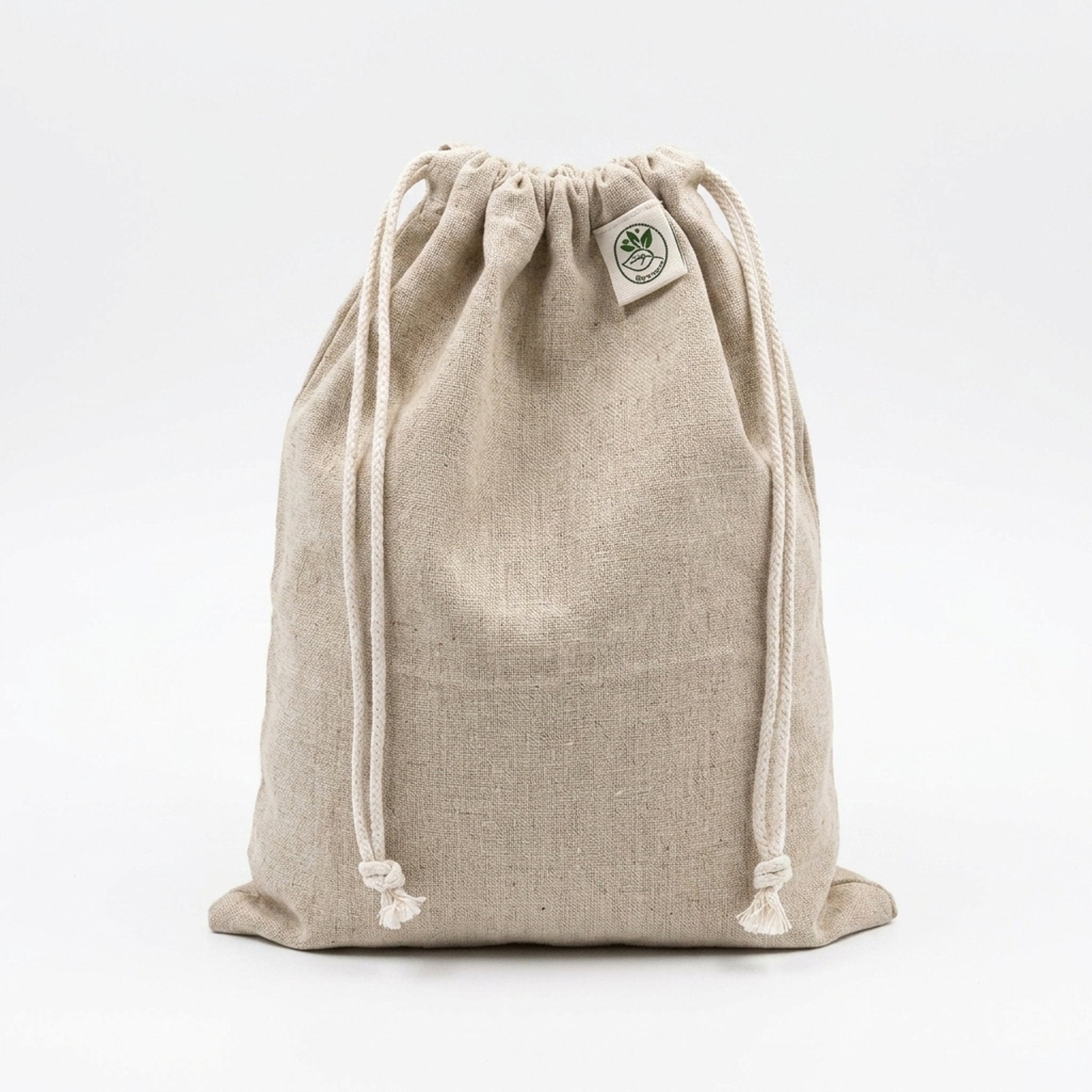 A natural beige hemp cotton drawstring bag with white drawstrings and a small circular green and white logo patch on the upper right side, photographed against a seamless white background.