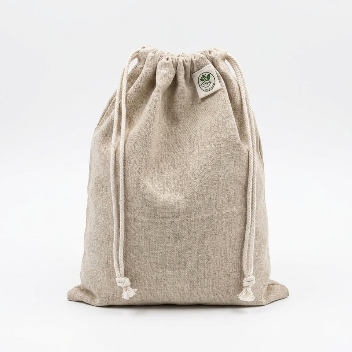 A natural beige hemp cotton drawstring bag with white drawstrings and a small circular green and white logo patch on the upper right side, photographed against a seamless white background.