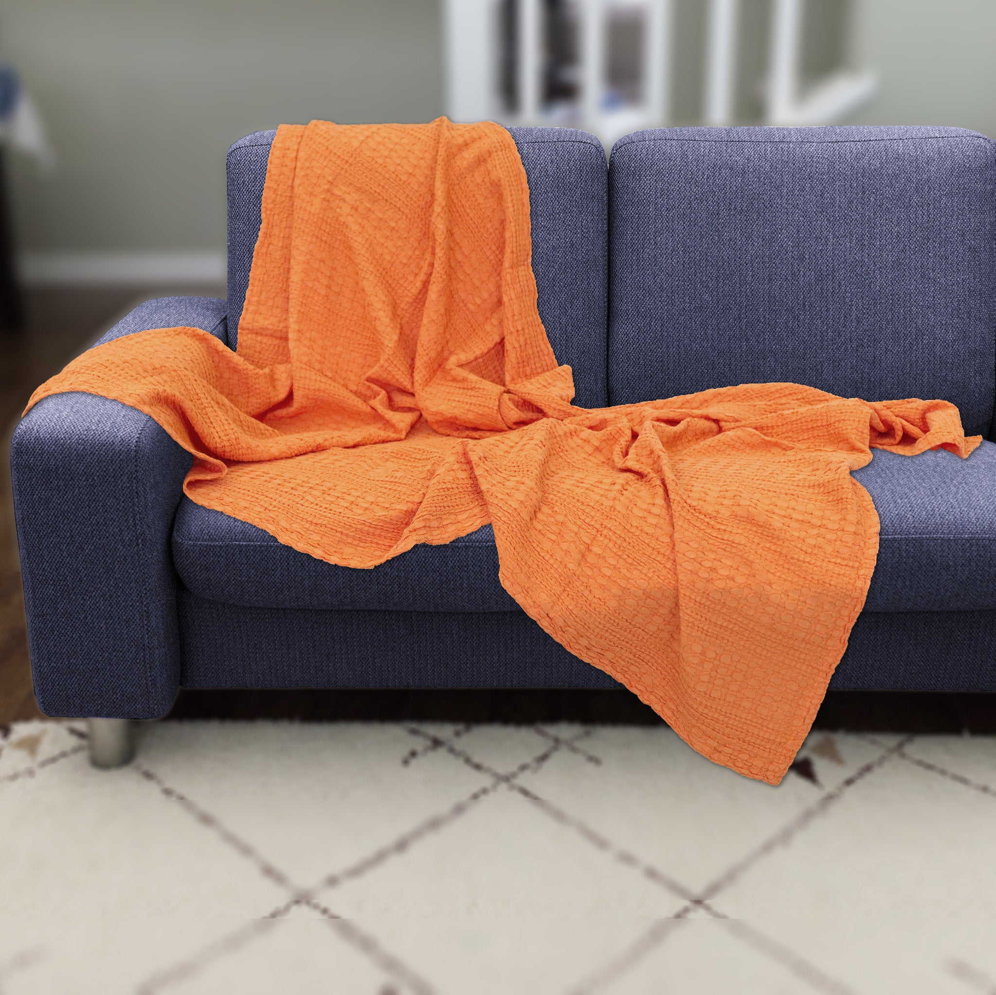 Orange textured wearable cotton throw of complex dual waffle weave from Hem And Tassel, draped over a blue sofa in a room with a tiled floor.