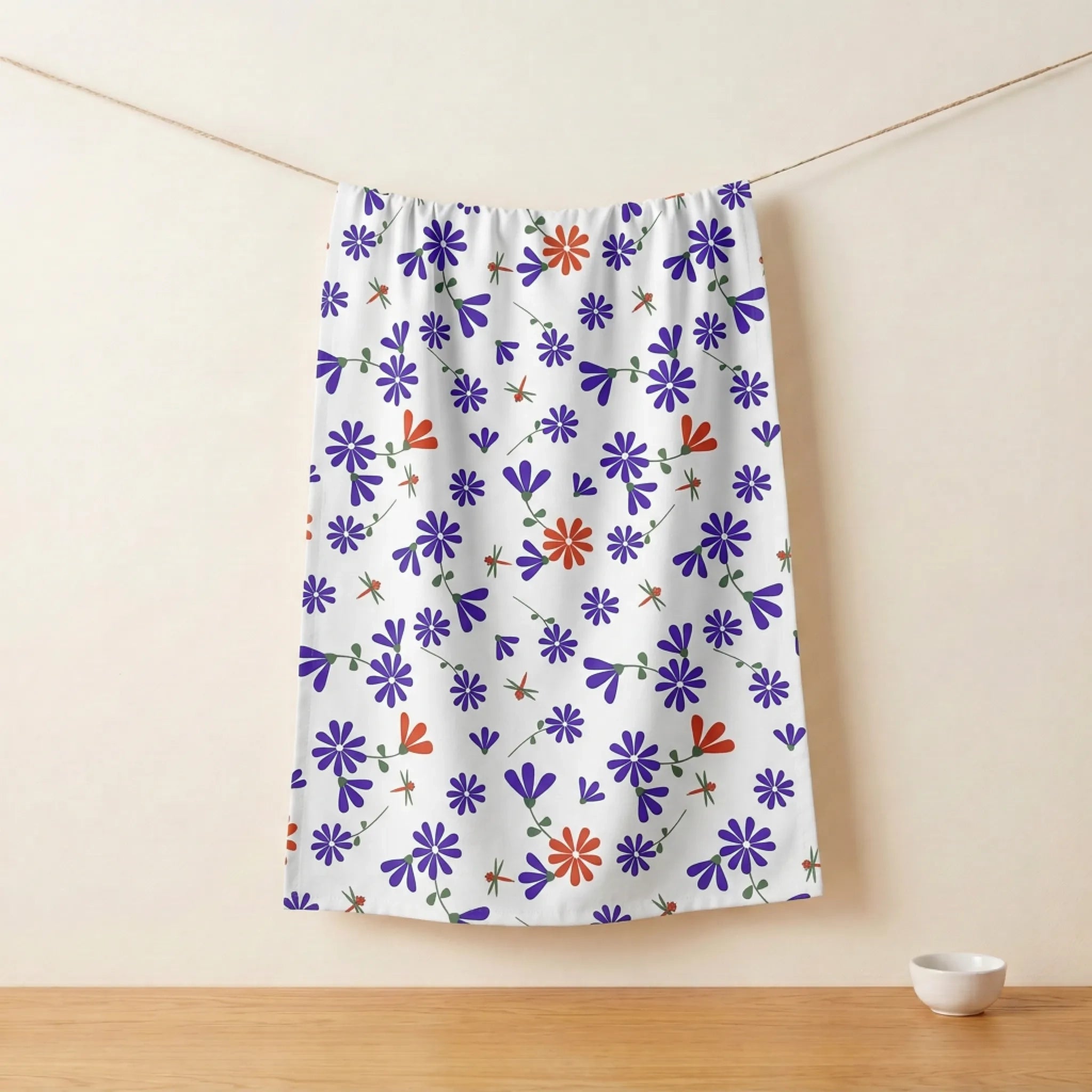 A white kitchen towel featuring a colorful pattern of purple and orange flowers, hanging from a string on a wooden table against a beige wall from Hem And Tassel 