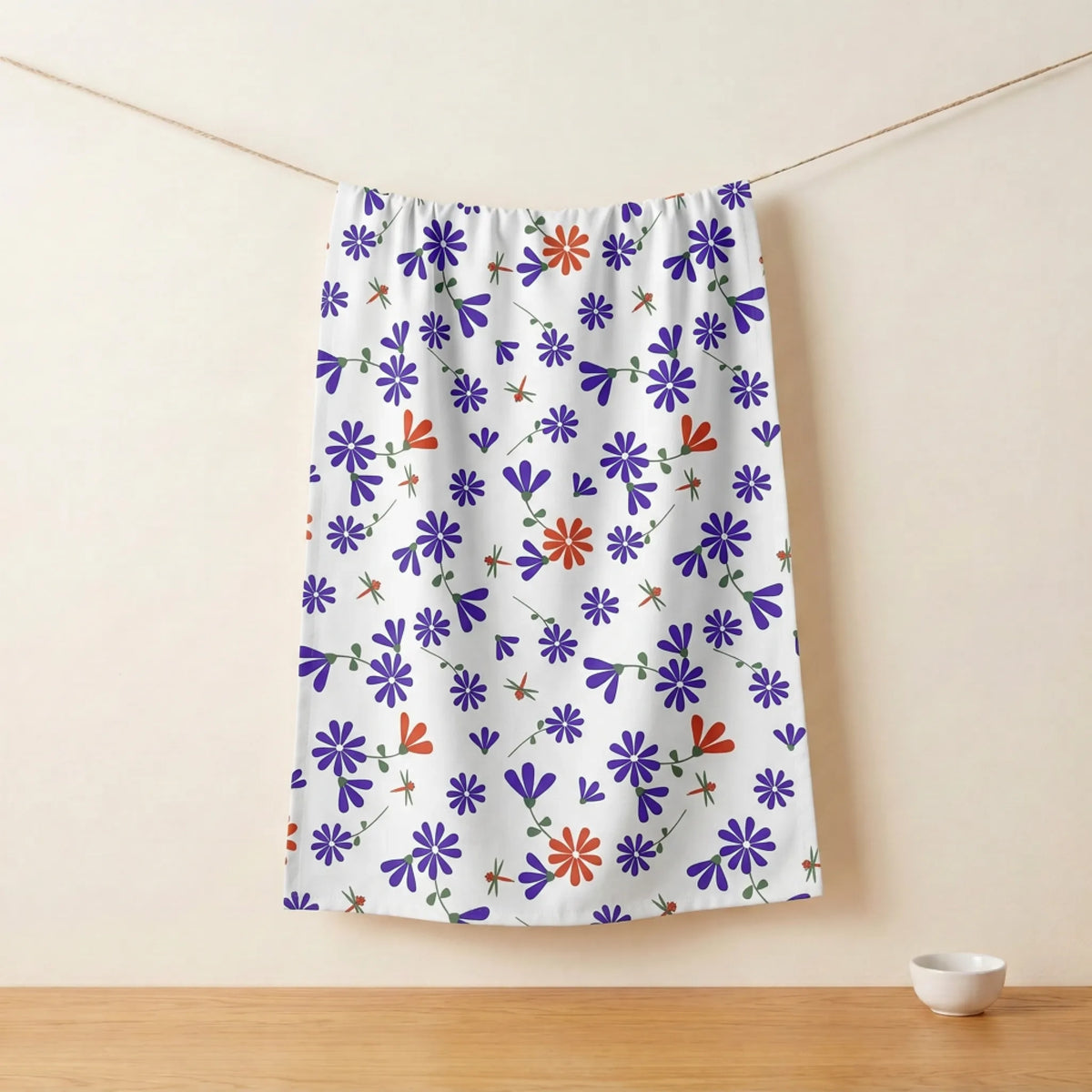 A white kitchen towel featuring a colorful pattern of purple and orange flowers, hanging from a string on a wooden table against a beige wall from Hem And Tassel 