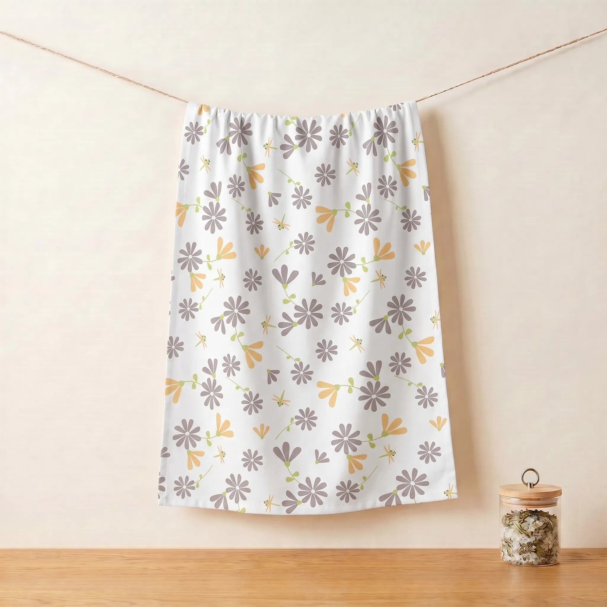 A white cotton kitchen towel with a repeating floral pattern in gray and buff hangs from a string on a wooden table against a beige wall, with a small glass jar on the right from Hem And Tassel 