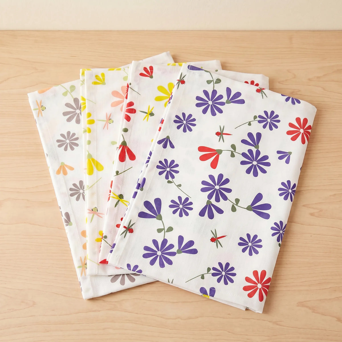 Four folded white kitchen towels with colorful floral patterns, arranged in a fan shape on a light wood surface from Hem And Tassel 
