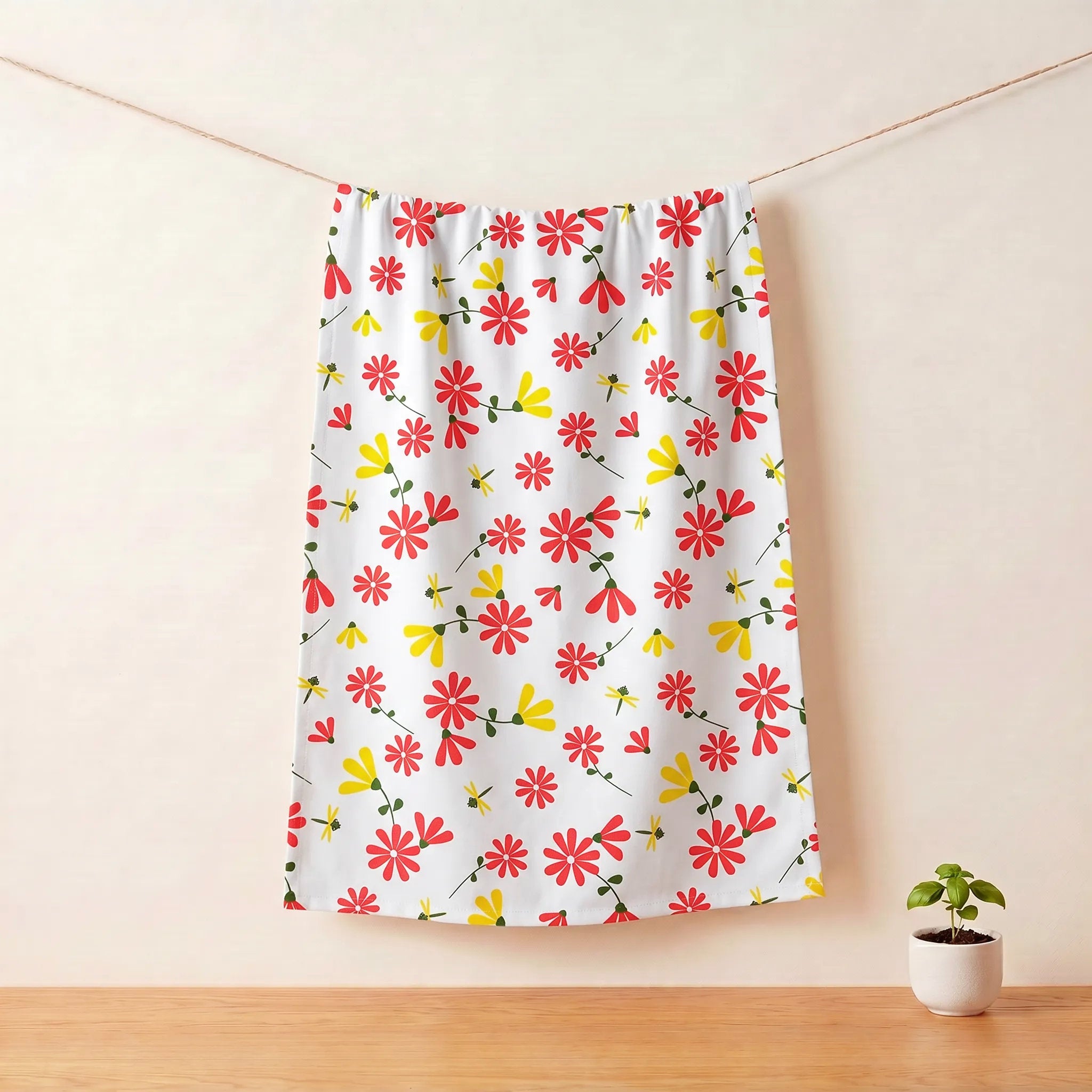 A white cotton kitchen towel with a colorful pattern of red, yellow, and orange flowers hanging on a clothesline in a minimalist room setting with a small potted plant on a wooden table from Hem And Tassel 