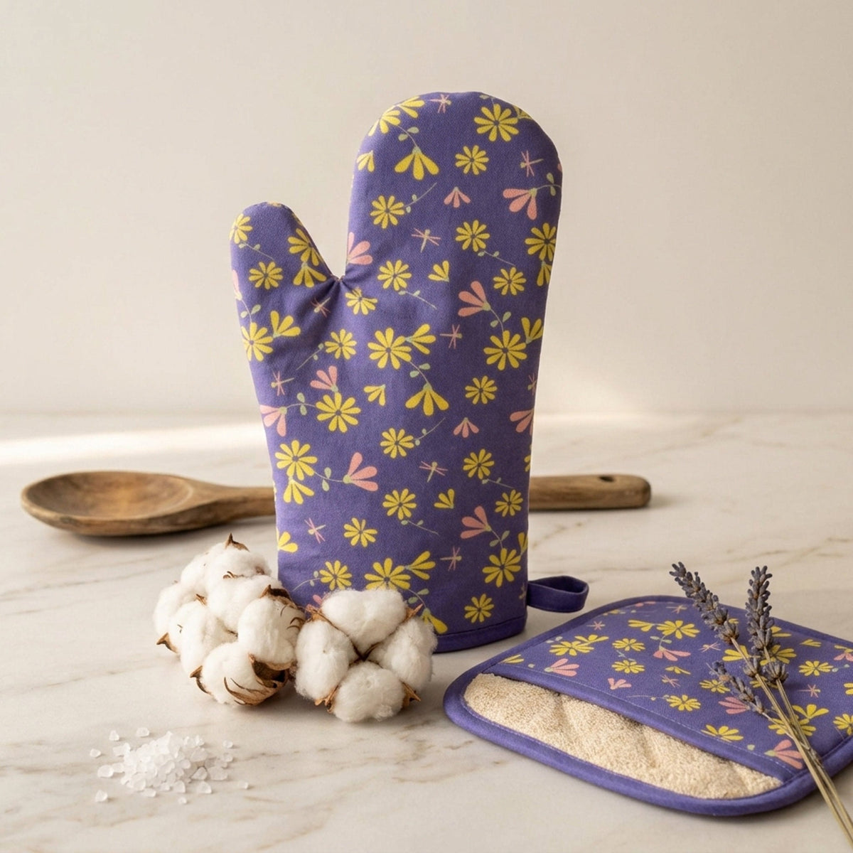 A purple oven mitt and matching pot holder with a yellow and pink floral print of the shy dragonfly collection, styled on a marble surface with cotton bolls, a wooden spoon, and lavender sprigs from Hem And Tassel
