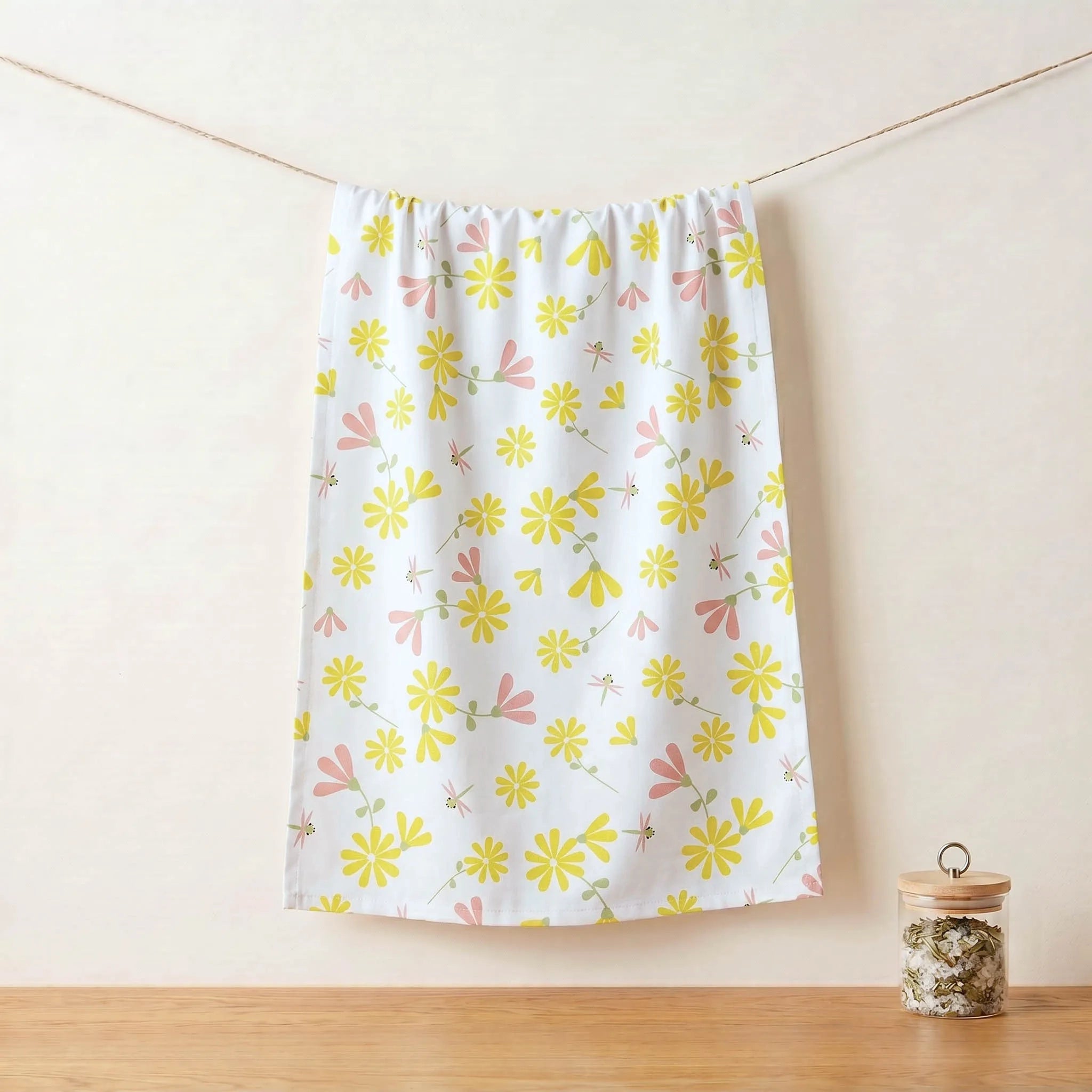 A white cotton kitchen towel with a colorful print of yellow flowers, pink blossoms, and green leaves, suspended from a string against a light beige wall above a wooden surface with a small jar from Hem And Tassel 