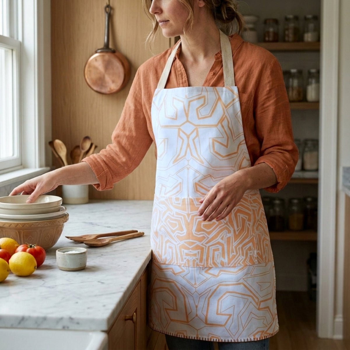 A woman wearing a white apron with a buff maze-like geometric pattern stands in a warm, rustic kitchen, reaching for bowls on a marble countertop from Hem And Tassel 