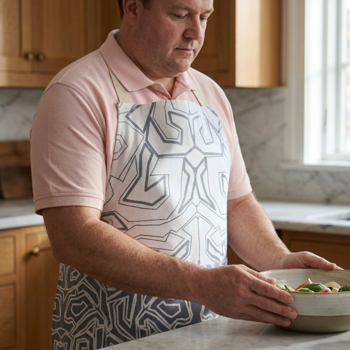 A man in a pink polo shirt wearing a white apron with a oil grey maze-like geometric pattern while preparing food in a kitchen.