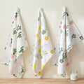 Three white kitchen towels with colorful cactus and pot patterns hang on hooks against a beige wall above a wooden surface, with a small glass jar on the right from Hem And Tassel 