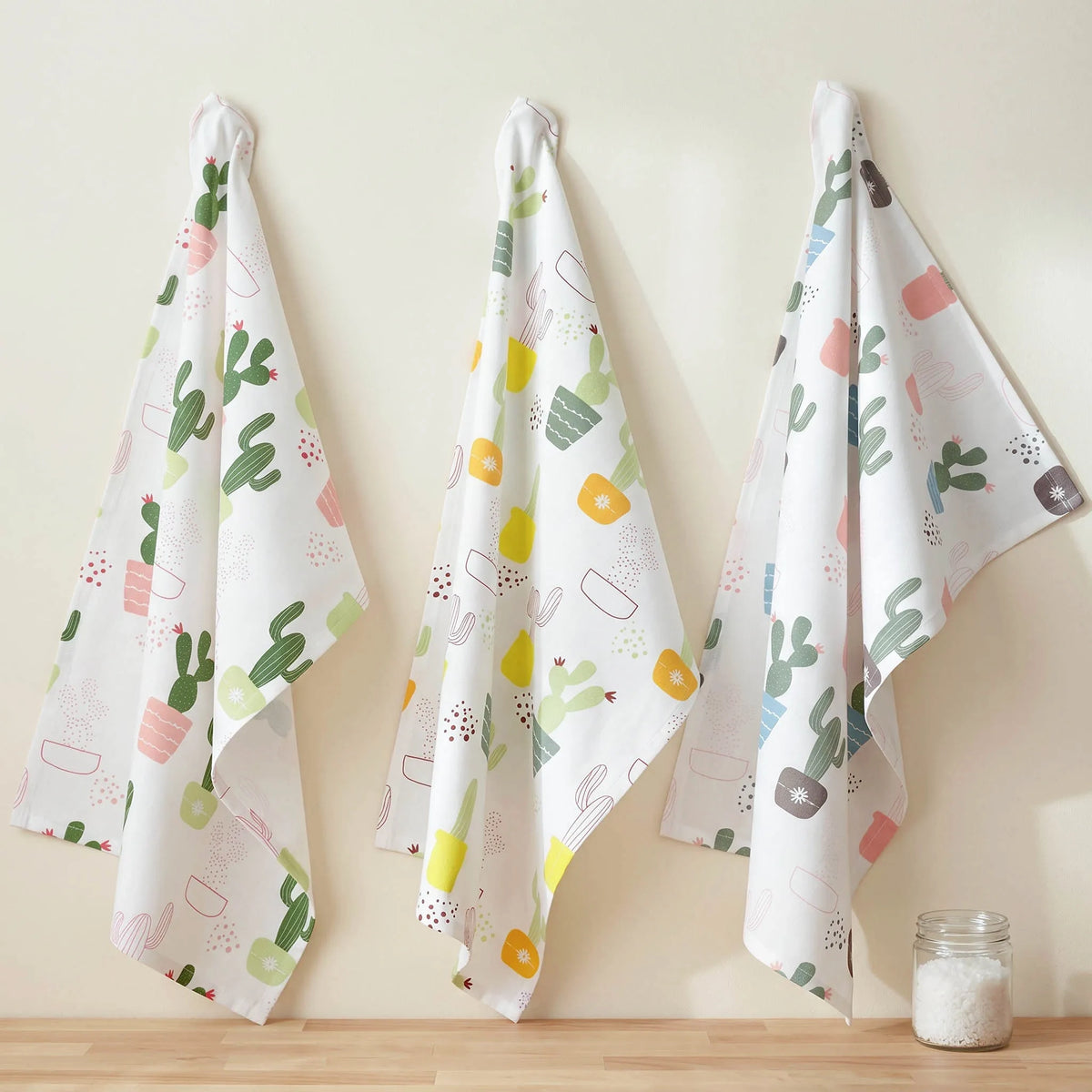 Three white kitchen towels with colorful cactus and pot patterns hang on hooks against a beige wall above a wooden surface, with a small glass jar on the right from Hem And Tassel 