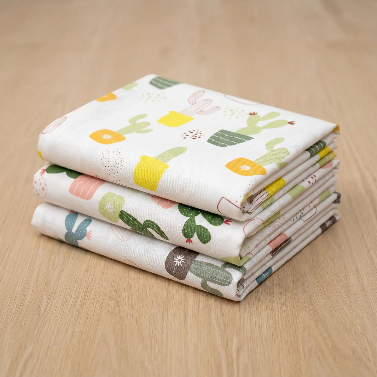 A stack of three folded white cotton kitchen towels, printed with a colorful cactus and succulent pattern, displayed on a light wood surface from Hem And Tassel 