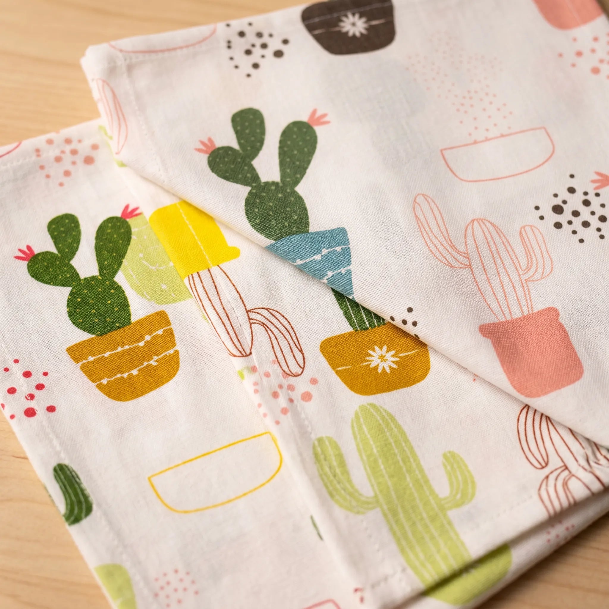three folded white fabric cotton kitchen towels with a colorful cactus and pot print, laid on a light wood surface from Hem And Tassel 