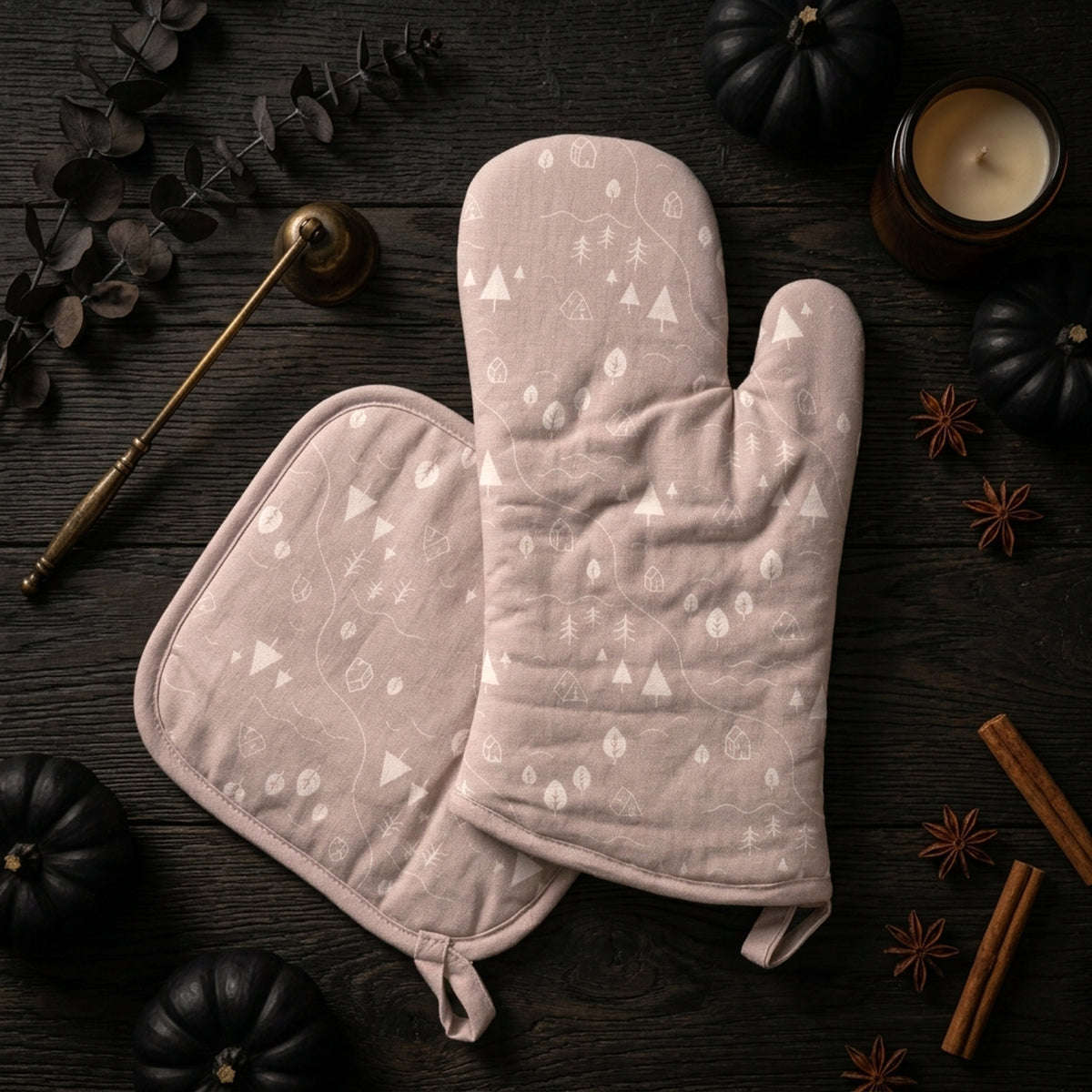 Two dusty beige oven mitts with a white forest pattern, arranged on a dark rustic wooden table surrounded by seasonal props like pumpkins, cinnamon sticks, star anise, and a brass candlestick from Hem And Tassel