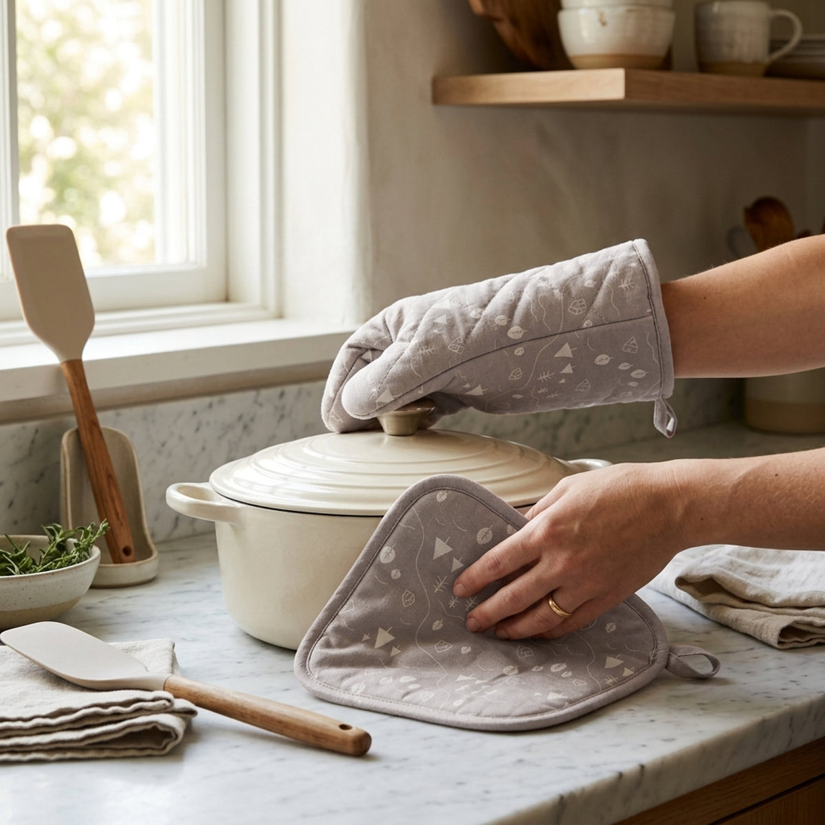 A person wearing quilted oven mitts with a light beige color and a tiny countryside pattern handles a cream-colored pot on a marble kitchen countertop from Hem And Tassel