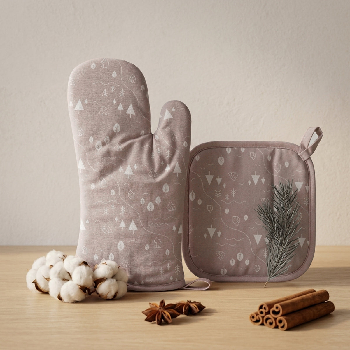 A matching oven mitt and pot holder set in a dusty mauve color with a white forest tree pattern, styled on a light wood surface with cotton bolls, star anise, cinnamon sticks, and a pine sprig against a neutral beige background from Hem And Tassel