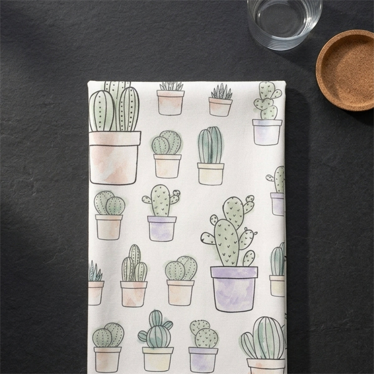 A rectangular white cotton kitchen towel , featuring a watercolor-style pattern of various cacti and succulents in pots, laid flat on a dark slate surface with a glass and a small wooden dish nearby from Hem And Tassel 