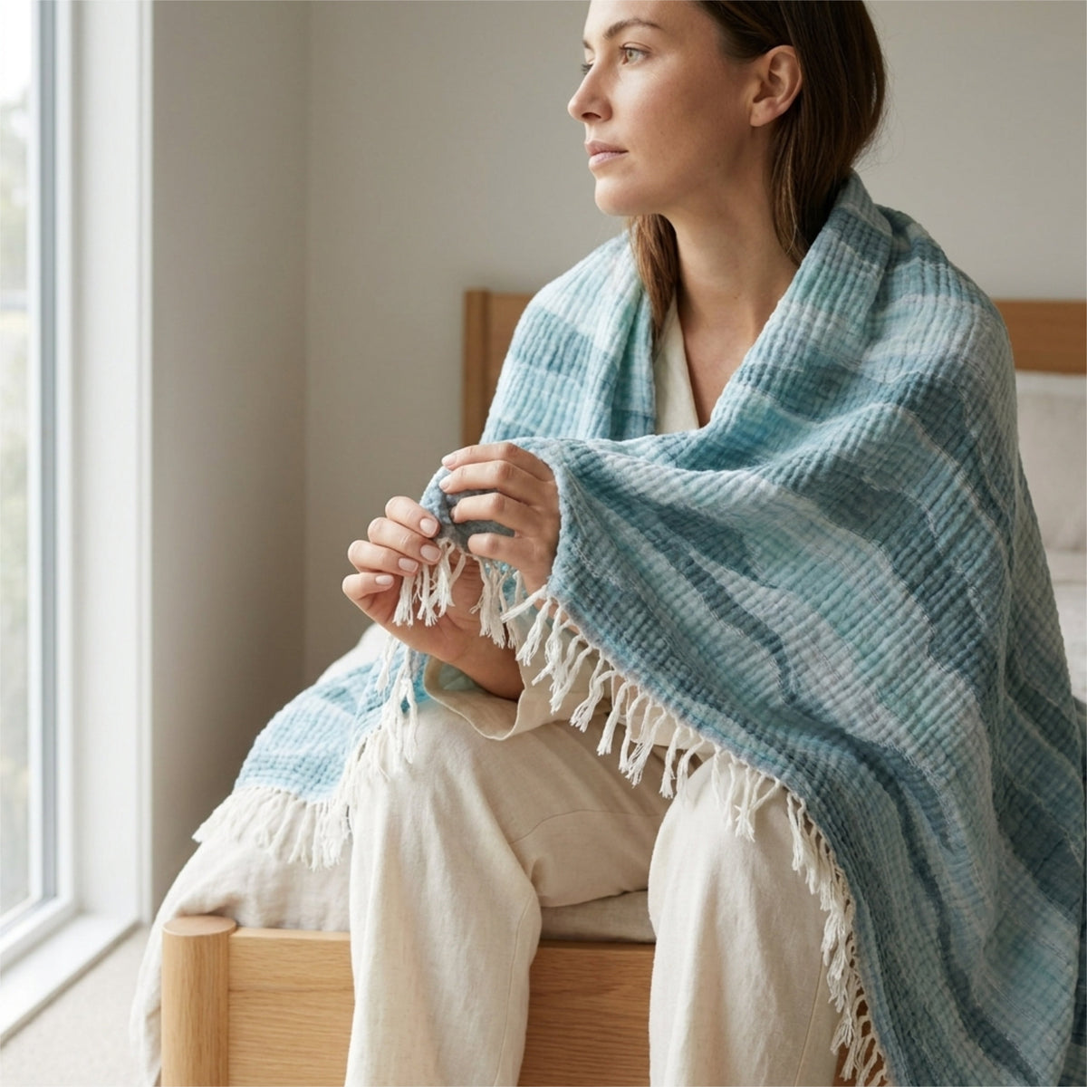A woman sits on a wooden bed in a softly lit room, wrapped in a seersucker muslin throw blanket with a waves pattern in three shades of blue, and white tassels. She is wearing cream-colored pants and holding a small blue cup from Hem And Tassel 