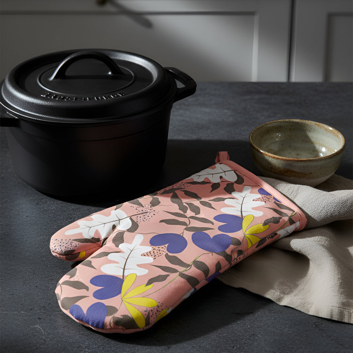 A floral oven mitt with a pink base and purple, yellow, and white leaves pattern rests on a dark countertop next to a black Dutch oven and a small ceramic bowl from Hem And Tassel