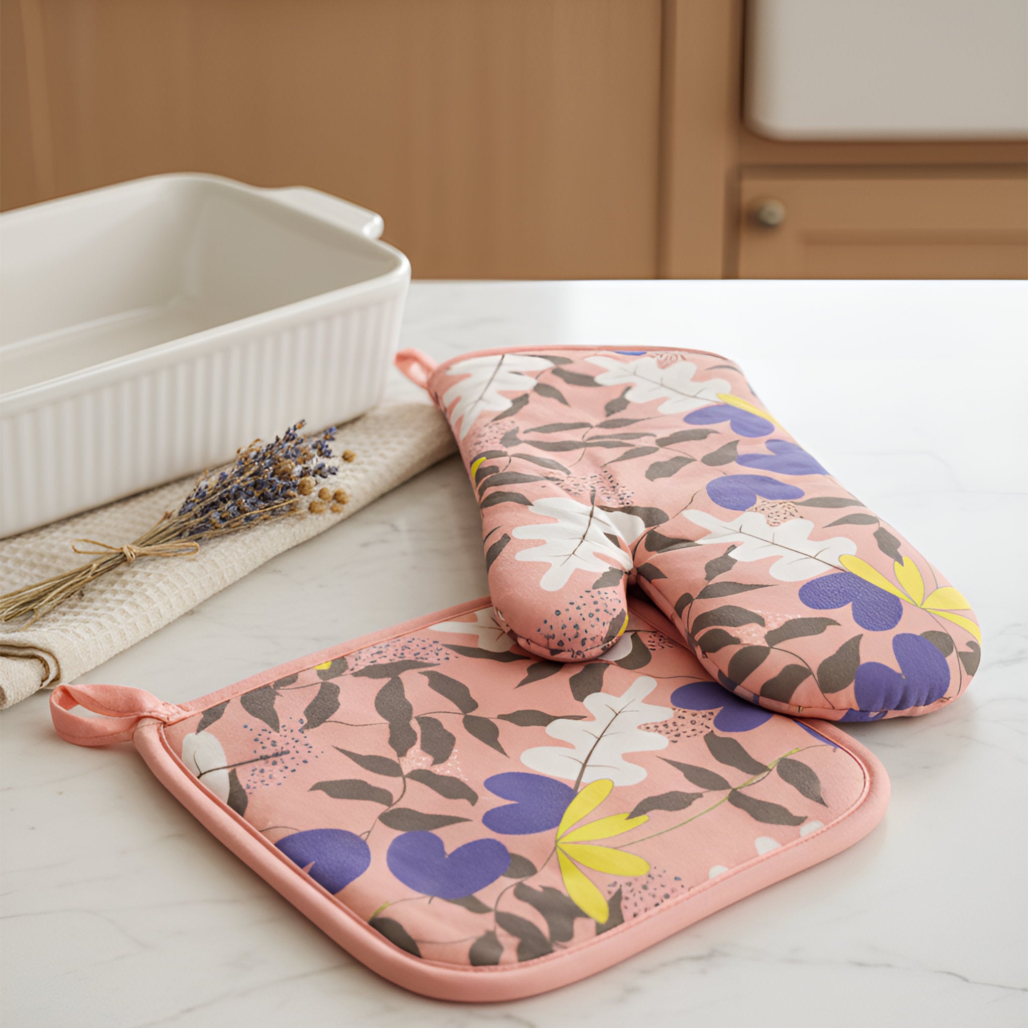A set of three kitchen oven accessories on a white marble countertop: a square potholder with a pink border and floral print, a matching rectangular oven mitt, and a white ceramic baking dish in the background. from Hem And Tassel 
