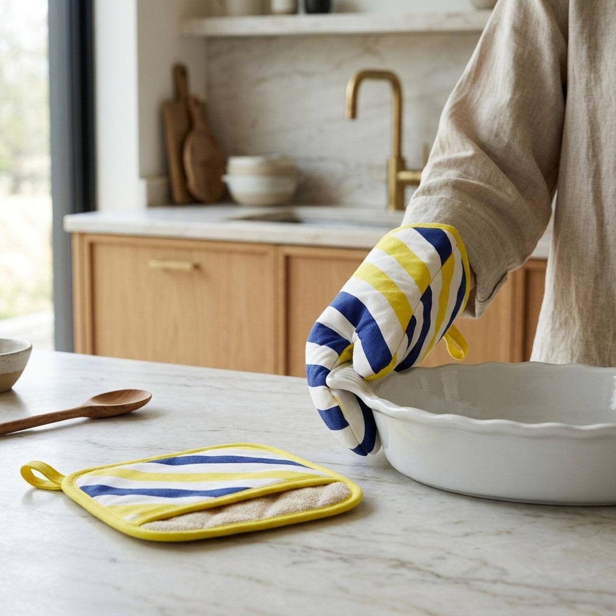 a woman handling a bowl wearing an oven mitt with navy and yellow stripes of the yellow paino collection while the matching  pot holder with Terry lining sits on a marble counter in the kitchen from Hem And Tassel