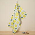 A white cotton kitchen towel with a yellow lemon and green leaf pattern, hanging from a wall hook above a wooden surface with a small ceramic peeler nearby from Hem And Tassel