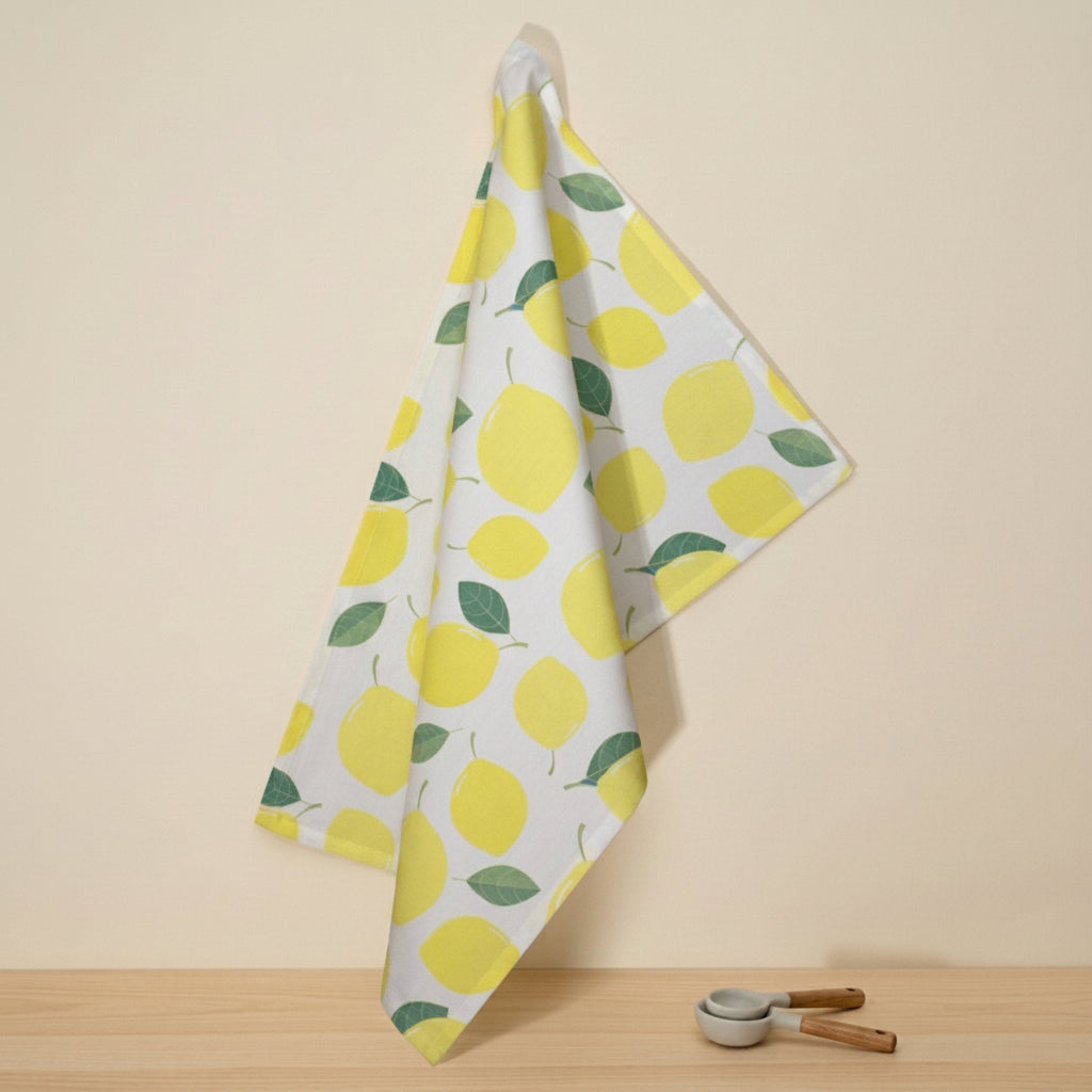 A white cotton kitchen towel with a yellow lemon and green leaf pattern, hanging from a wall hook above a wooden surface with a small ceramic peeler nearby from Hem And Tassel
