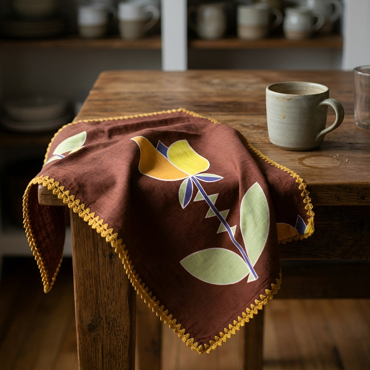 A brown cotton dinner napkin with a yellow and green floral pattern and gold tassel trim, draped over a rustic wooden table next to a ceramic mug from Hem And Tassel