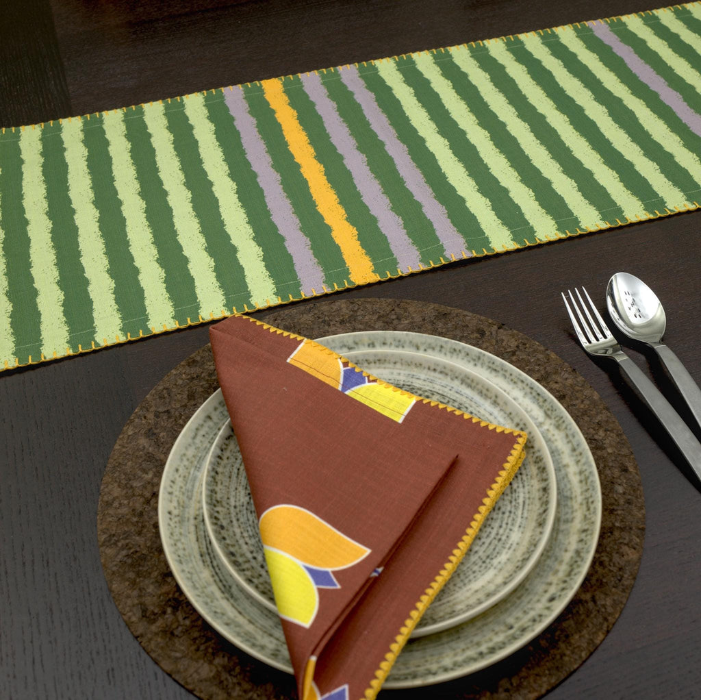 Dining table setting with a striped runner, folded art deco lip tulip dinner napkin and cutlery on a dark surface from hem and tassel