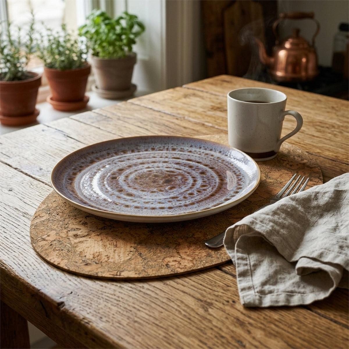 A round ceramic plate with concentric textured rings in earthy brown, beige, and muted blue tones, styled on a rustic wooden table with a graphic bark peel cork placemat, a white mug, a linen napkin, and potted plants in the background from Hem And Tassel 