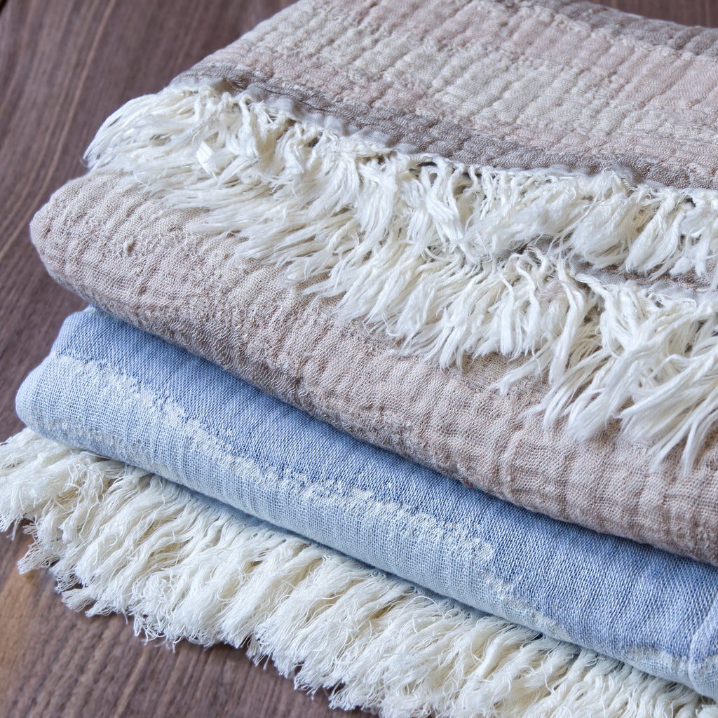 Stack of beige abd blue seersucker 'waves' wearable throw blankets with fringes on a wooden surface
