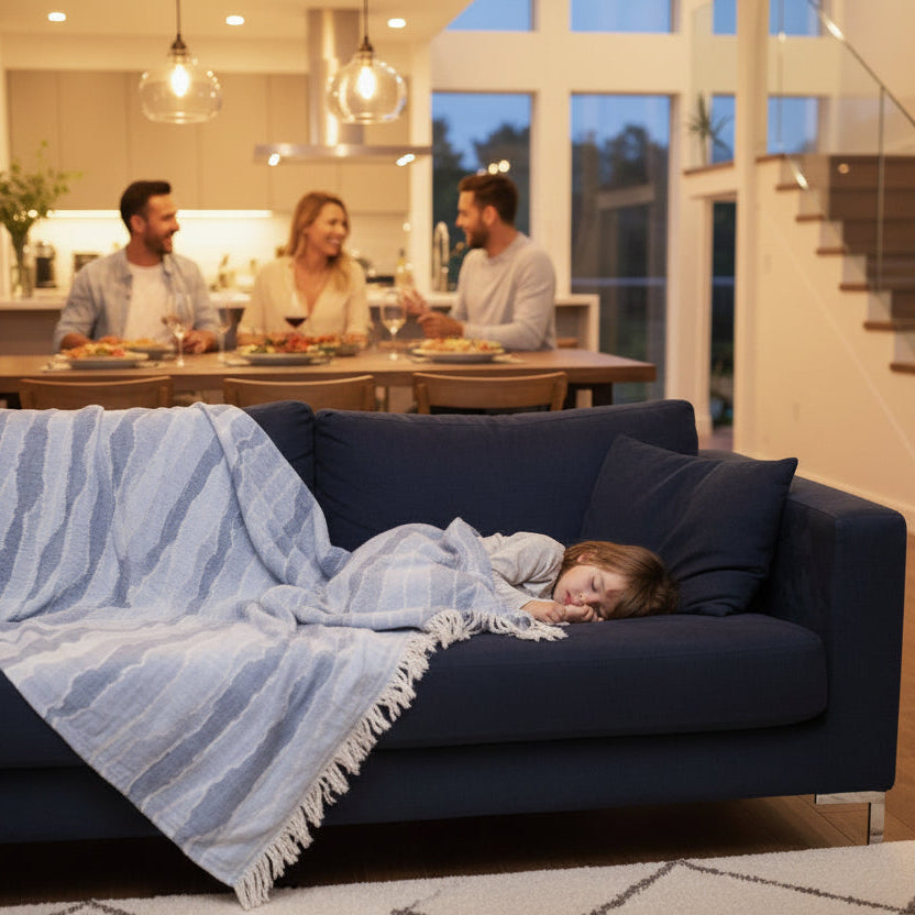 Child lying on a blue couch with a striped waves pattern throw blanket, while people are dining in the background.