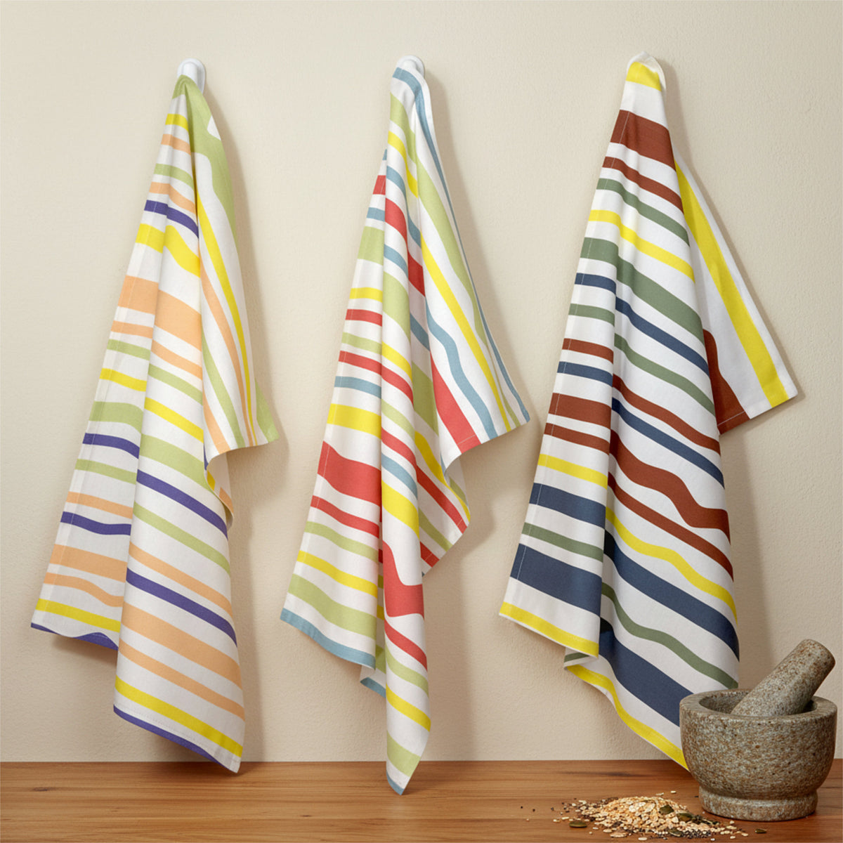 broad-width stripes in Spring, summer and Fall colors on 3 cotton kitchen towels hanging on a beige wall with a mortar and pestle and some grains on a wood counter below from Hem And Tassel 