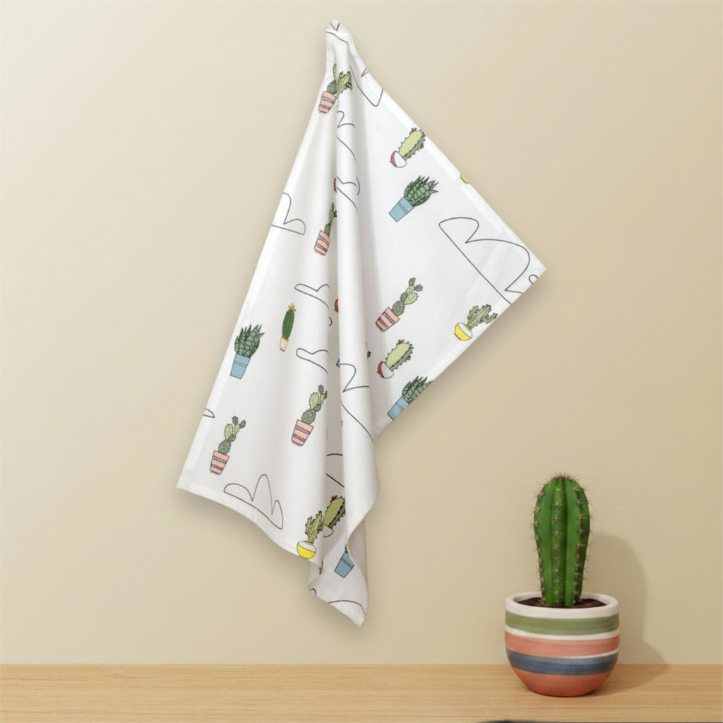 A white kitchen towel with a cactus and plant pattern hangs on a beige wall above a wooden surface, with a small potted cactus in a striped pot on the right from Hem And Tassel