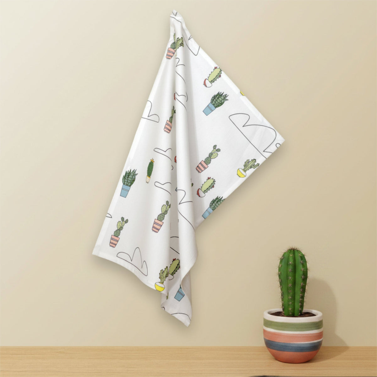 A white kitchen towel with a cactus and plant pattern hangs on a beige wall above a wooden surface, with a small potted cactus in a striped pot on the right from Hem And Tassel