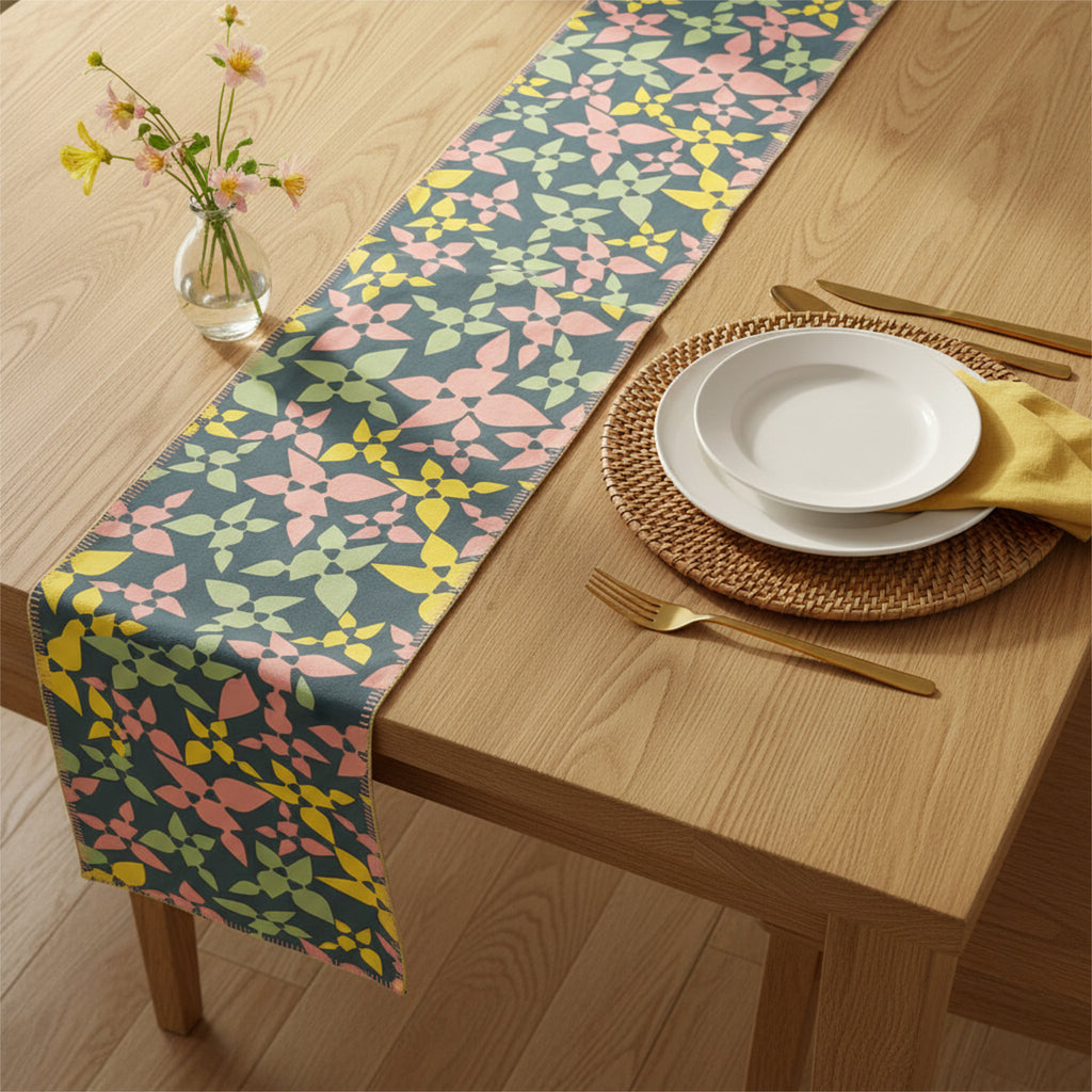 A floral table runner with a repeating geometric flower pattern in pink, yellow, and green on a slate blue-gray background, styled on a light wood dining table with plates, a napkin, cutlery, and a small vase of flowers.