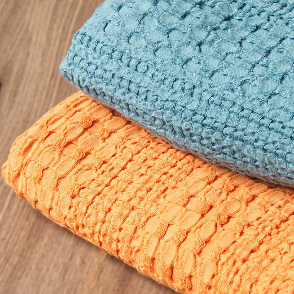 Closeup of two complex waffle wearable throws, one blue and one orange from Hem And Tassel , stacked on a wooden surface.