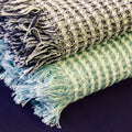 Two stonewashed waffle kitchen towels with fringes, one blue and white, the other green and white, on a dark background from Hem And Tassel 