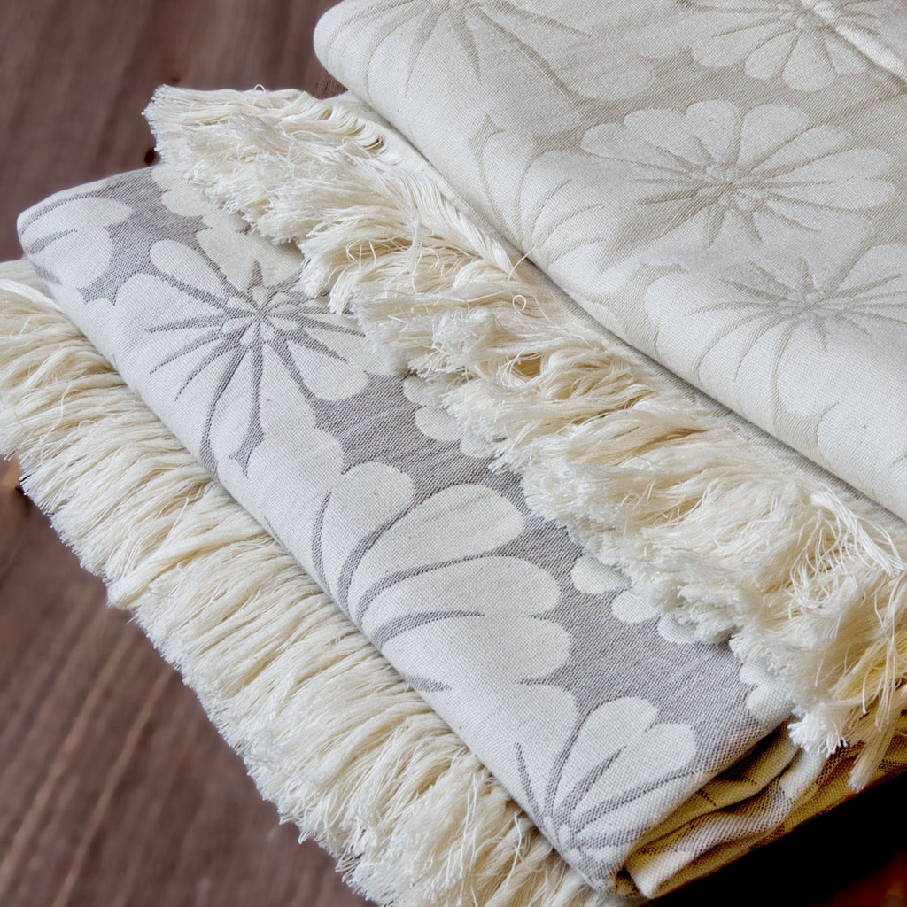 close up of wearable throws in pure cotton of bloom design in beige and slate colors, from Hem And Tassel, on a wood background