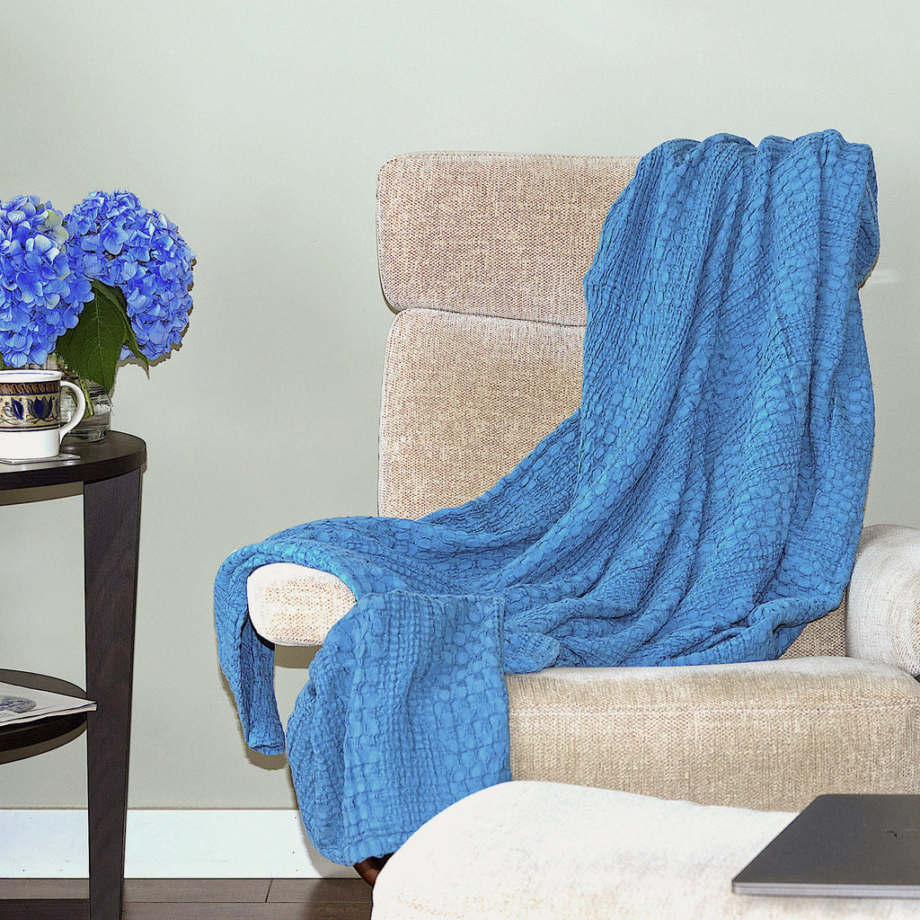 Blue textured wearable cotton throw of complex waffle weave from Hem And Tassel, draped over a beige armchair with a side table featuring blue flowers