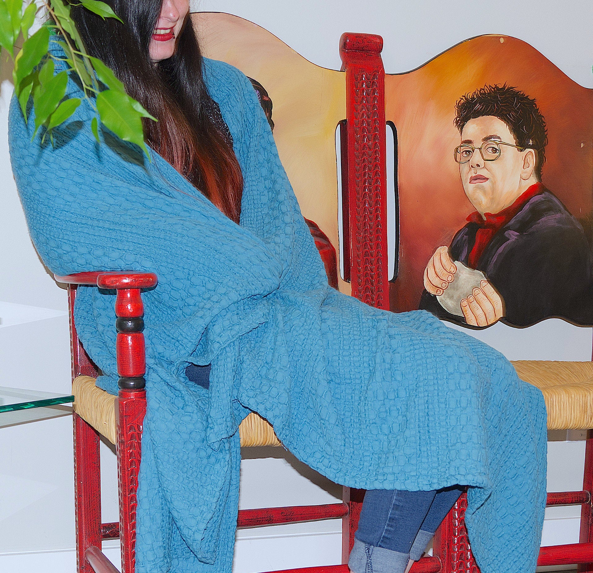 Person draped in a blue complex waffle wearable throw in cotton sitting on a red chair with a painting of a man holding a stone in the background.