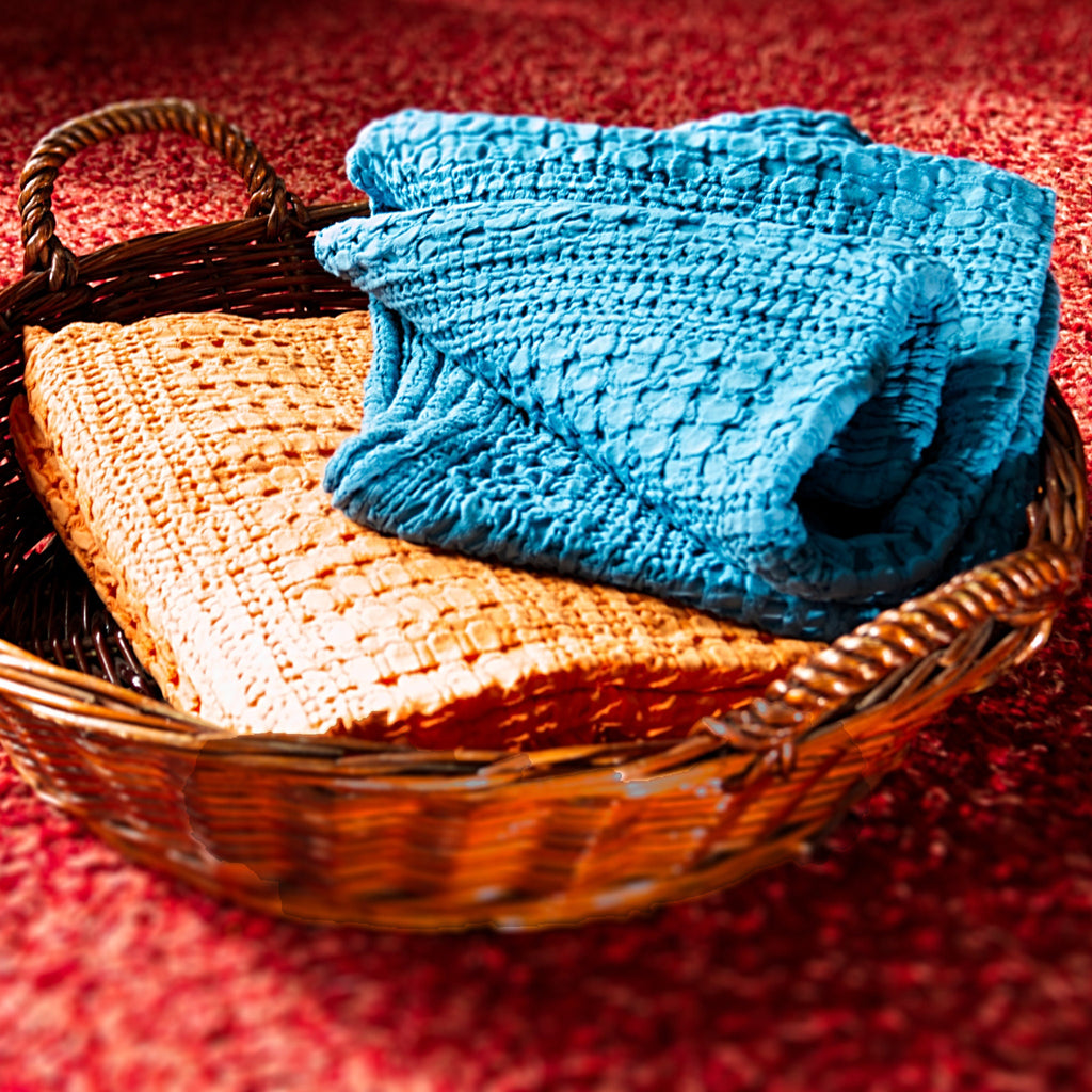 two wearable throws of complex waffle design in blue and orange from Hem And Tassel, in a Basket on a red textured surface