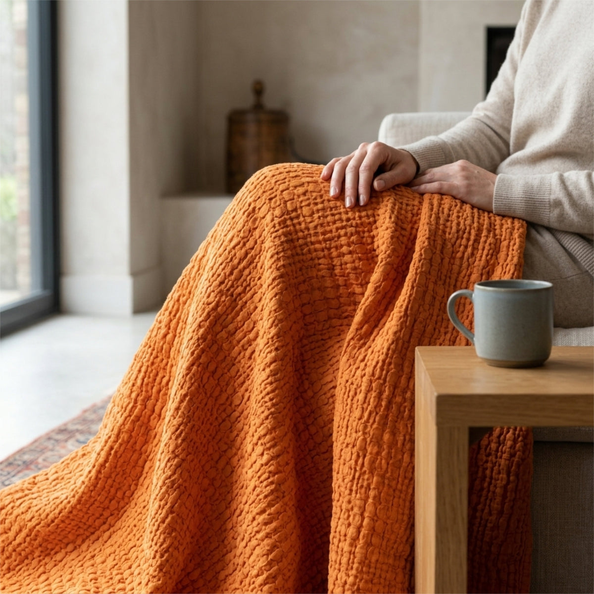 A pumpkin orange complex waffle throw blanket, hemmed on four sides is draped on the lap of a person with a coffee table and cup of coffee beside. from Hem And Tassel
