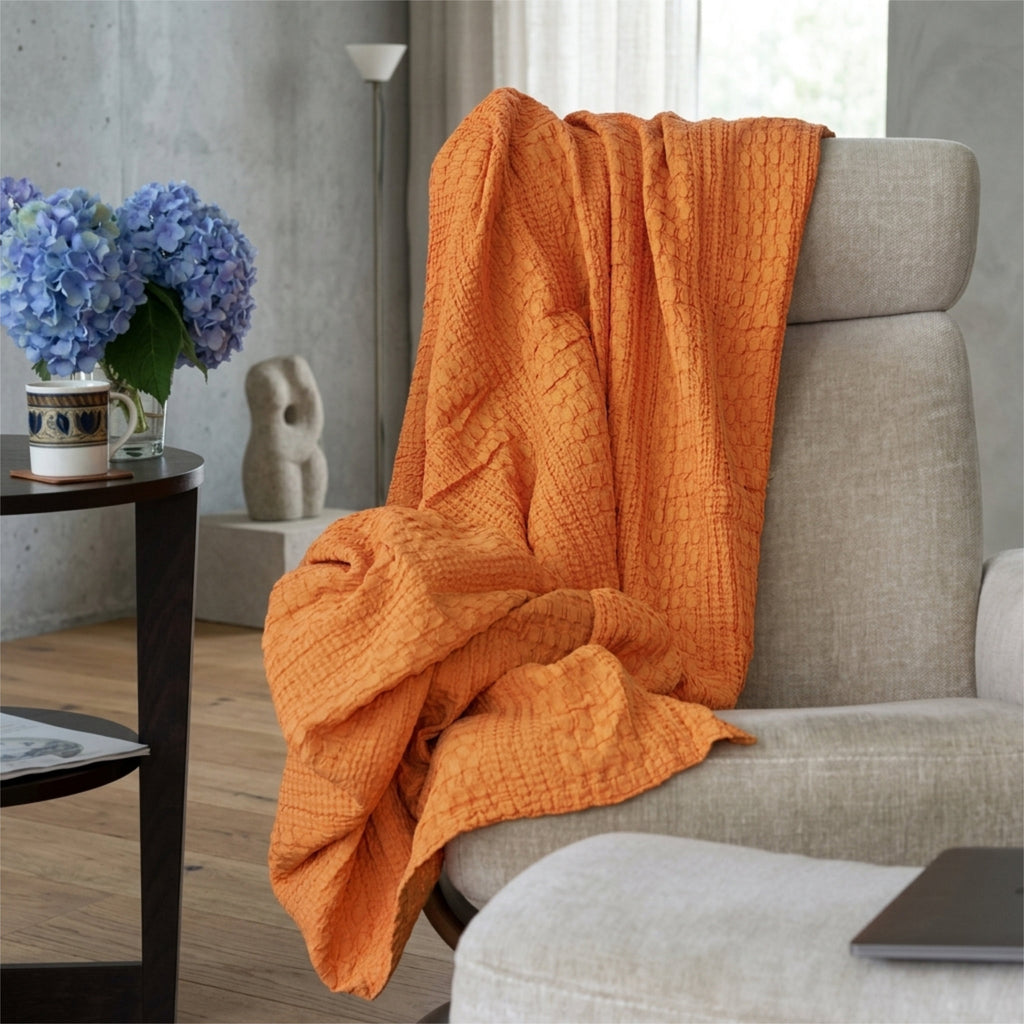 A pumpkin orange complex waffle throw blanket, hemmed on four sides, draped over a light gray upholstered armchair in a modern living room setting from Hem And Tassel 