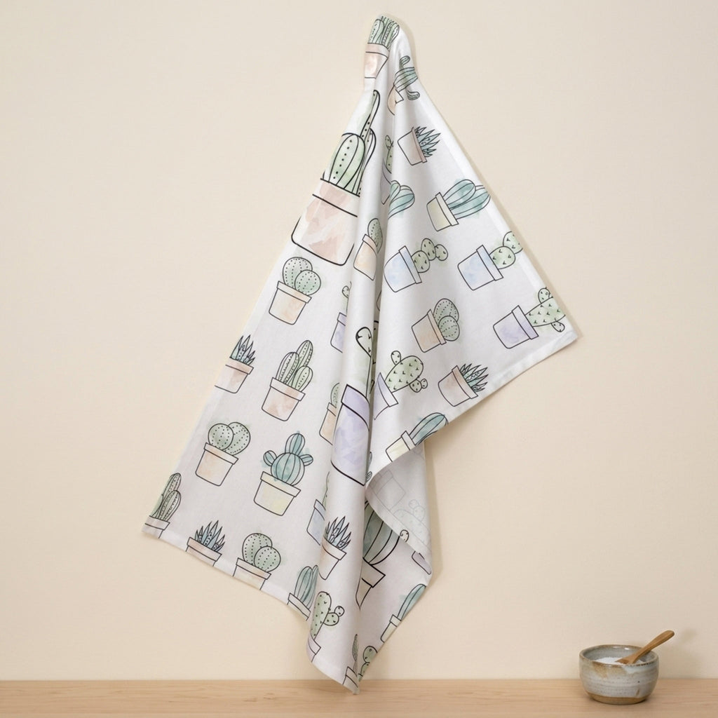 a cotton kitchen towel with an all-over watercolor print of several cacti in pots hangs on a wall above a wooden kitchen counter and small ceramic bowl with spoon in the salt sits below from Hem And Tassel 