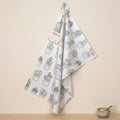 a cotton kitchen towel with an all-over watercolor print of several cacti in pots hangs on a wall above a wooden kitchen counter and small ceramic bowl with spoon in the salt sits below from Hem And Tassel 