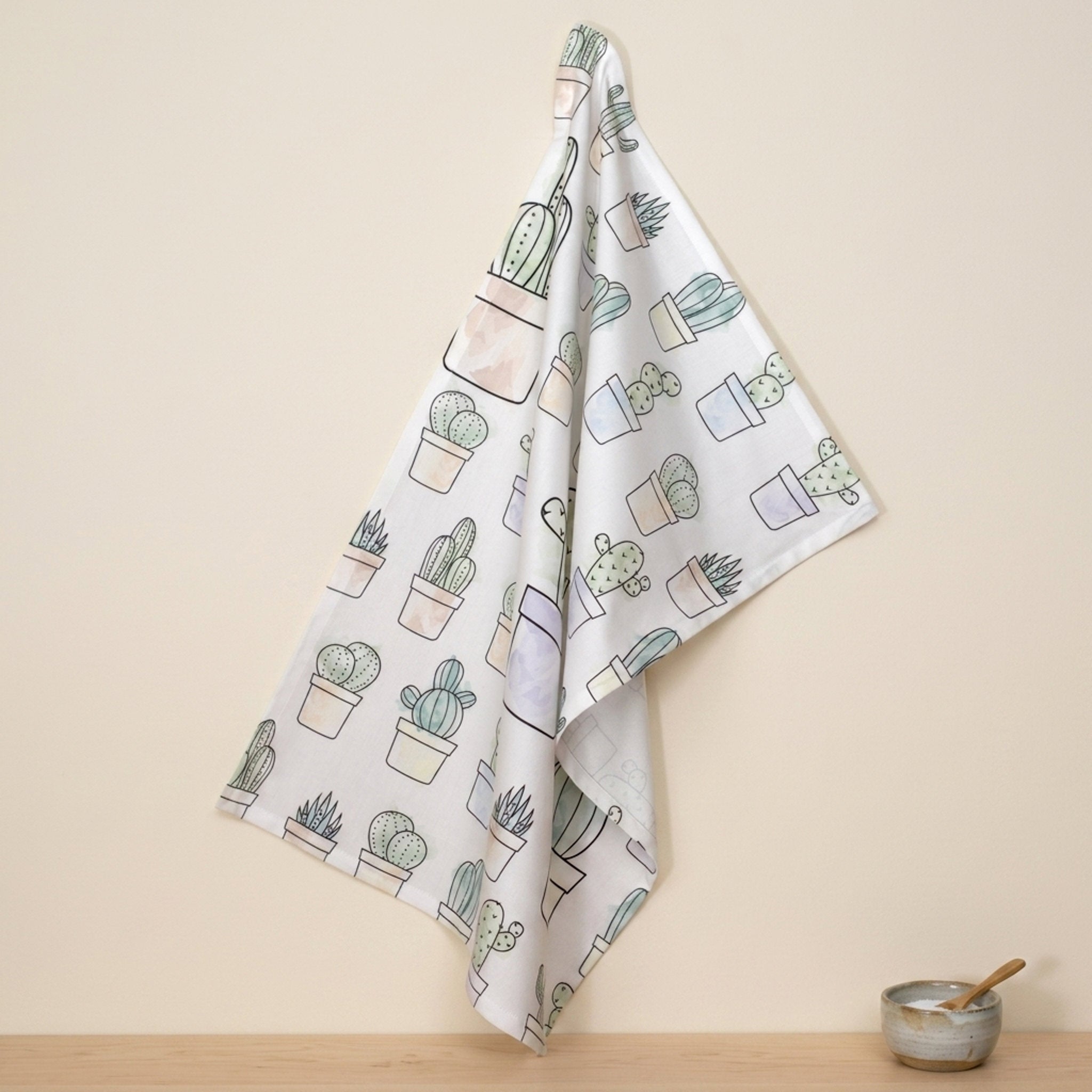 a cotton kitchen towel with an all-over watercolor print of several cacti in pots hangs on a wall above a wooden kitchen counter and small ceramic bowl with spoon in the salt sits below from Hem And Tassel 