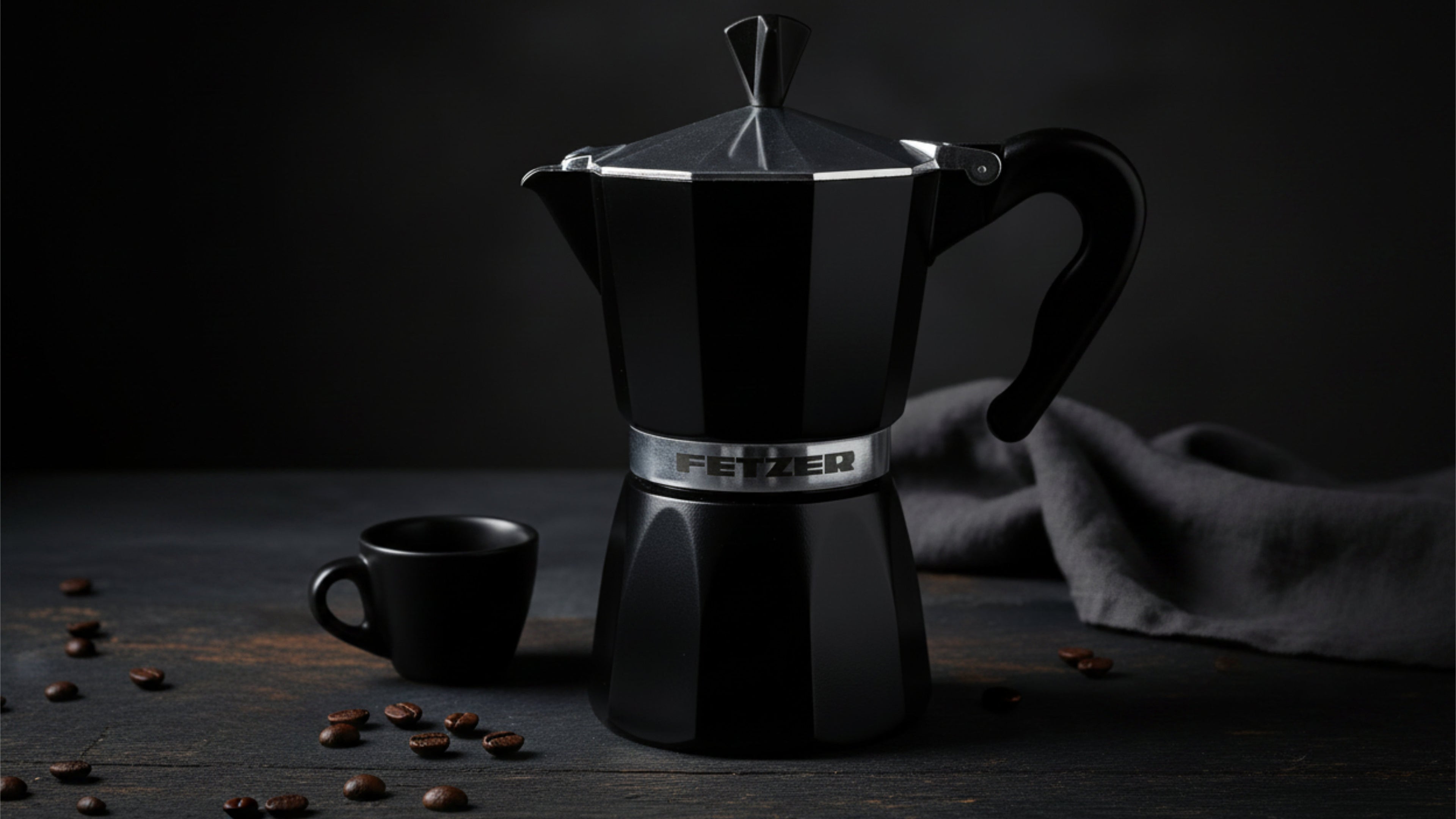 Black stovetop espresso maker with a cup and coffee beans on a dark surface