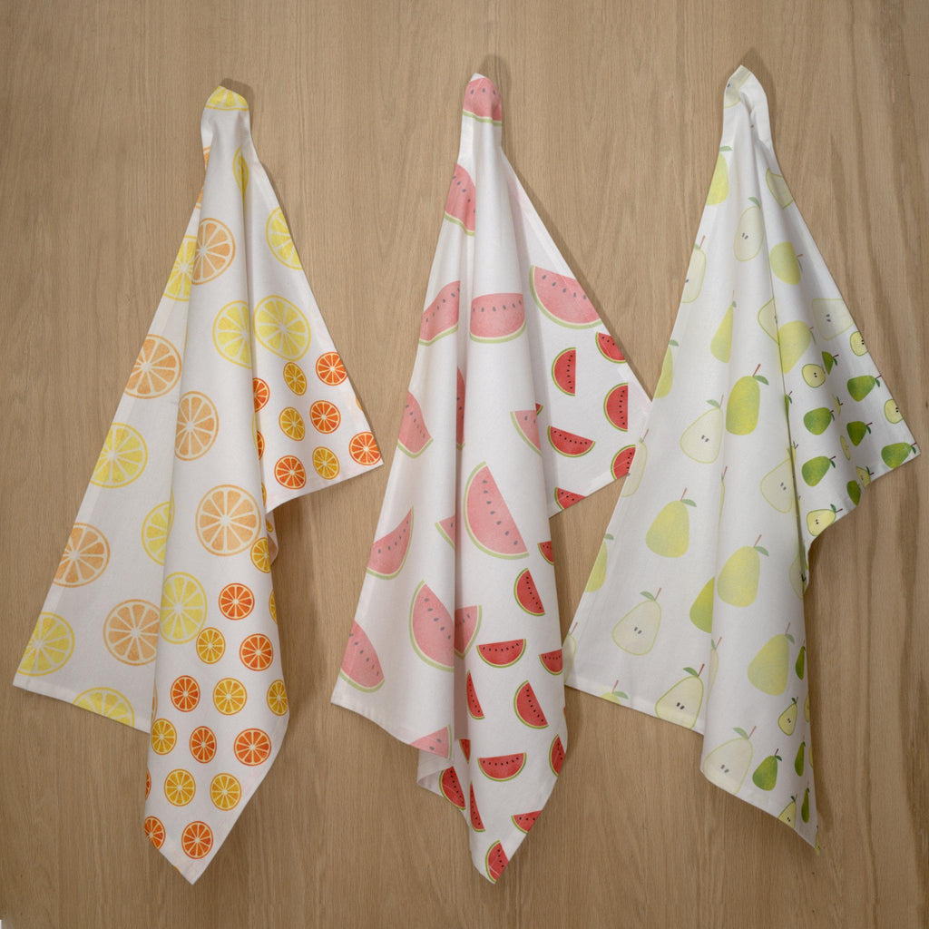 Three faded fruit kitchen towels with fruit patterns hanging on a wooden surface from Hem And Tassel 